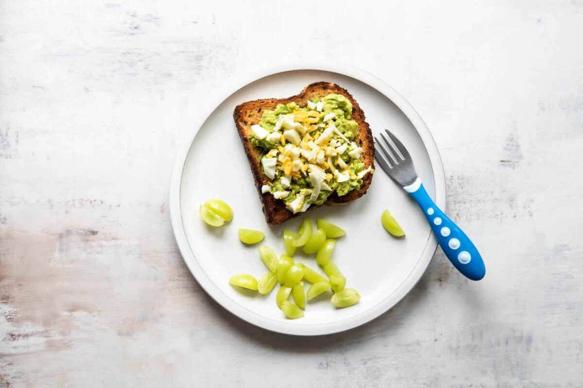 A plate with avocado toast topped with hard-boiled egg and grapes on the side.