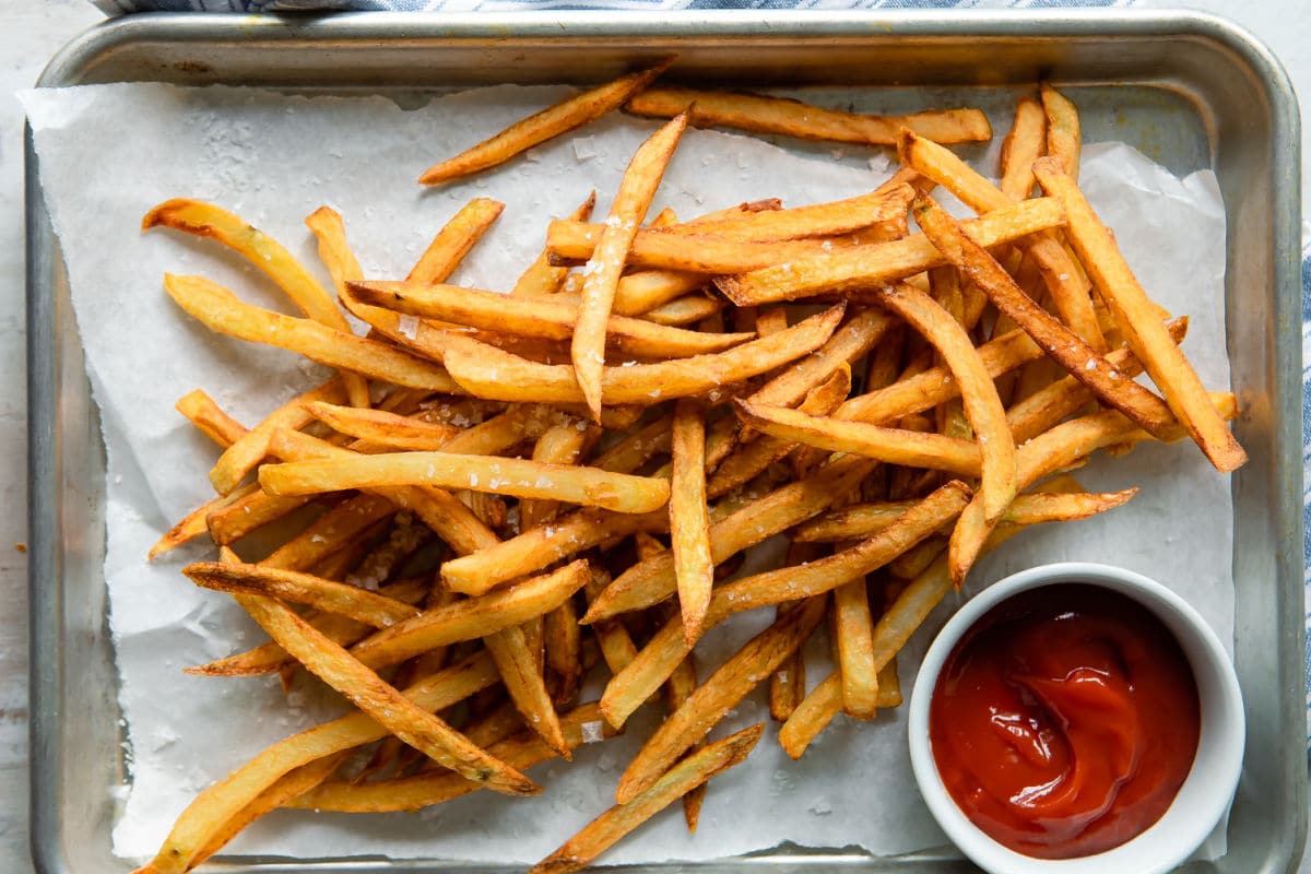 A pile of french fries on a baking sheet next to a cup of ketchup.