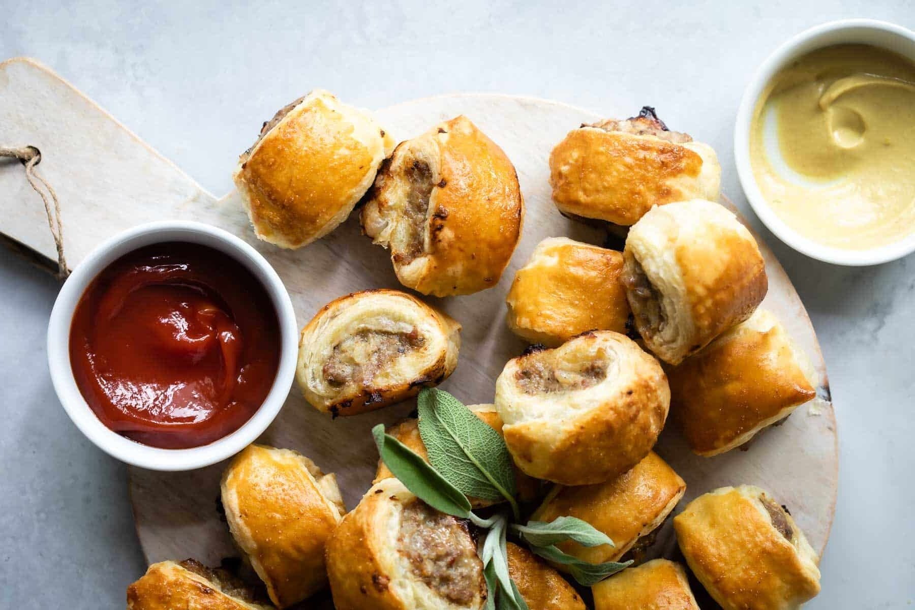 A wooden cutting board with sausage rolls next to a small cup of ketchup.