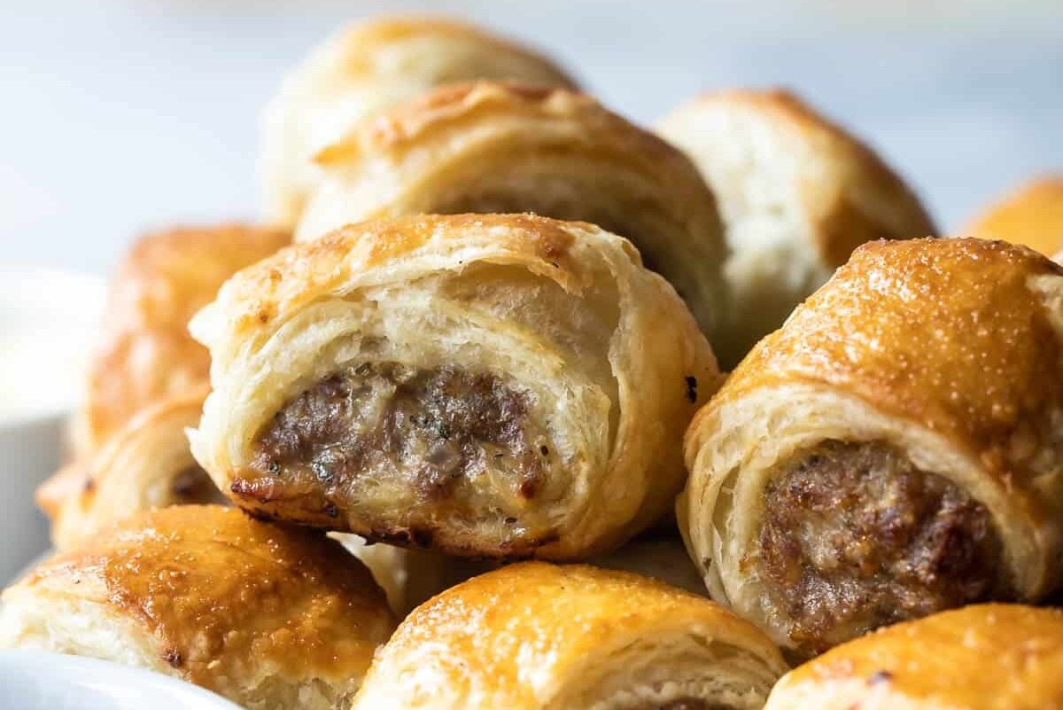 A wooden cutting board with sausage rolls next to a small cup of ketchup.