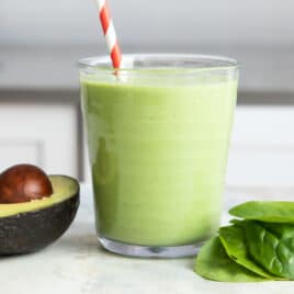A glass of avocado smoothie next to a pile of spinach and half of an avocado.