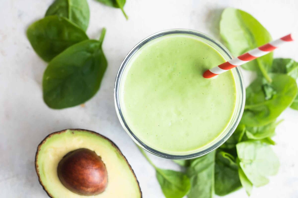 A glass of avocado smoothie next to a pile of spinach and half of an avocado.