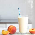 A glass of peach smoothie next to fresh peaches on a counter.