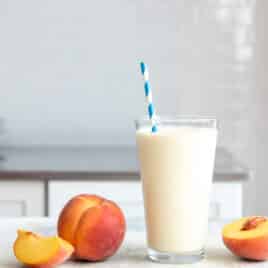 A glass of peach smoothie next to fresh peaches on a counter.