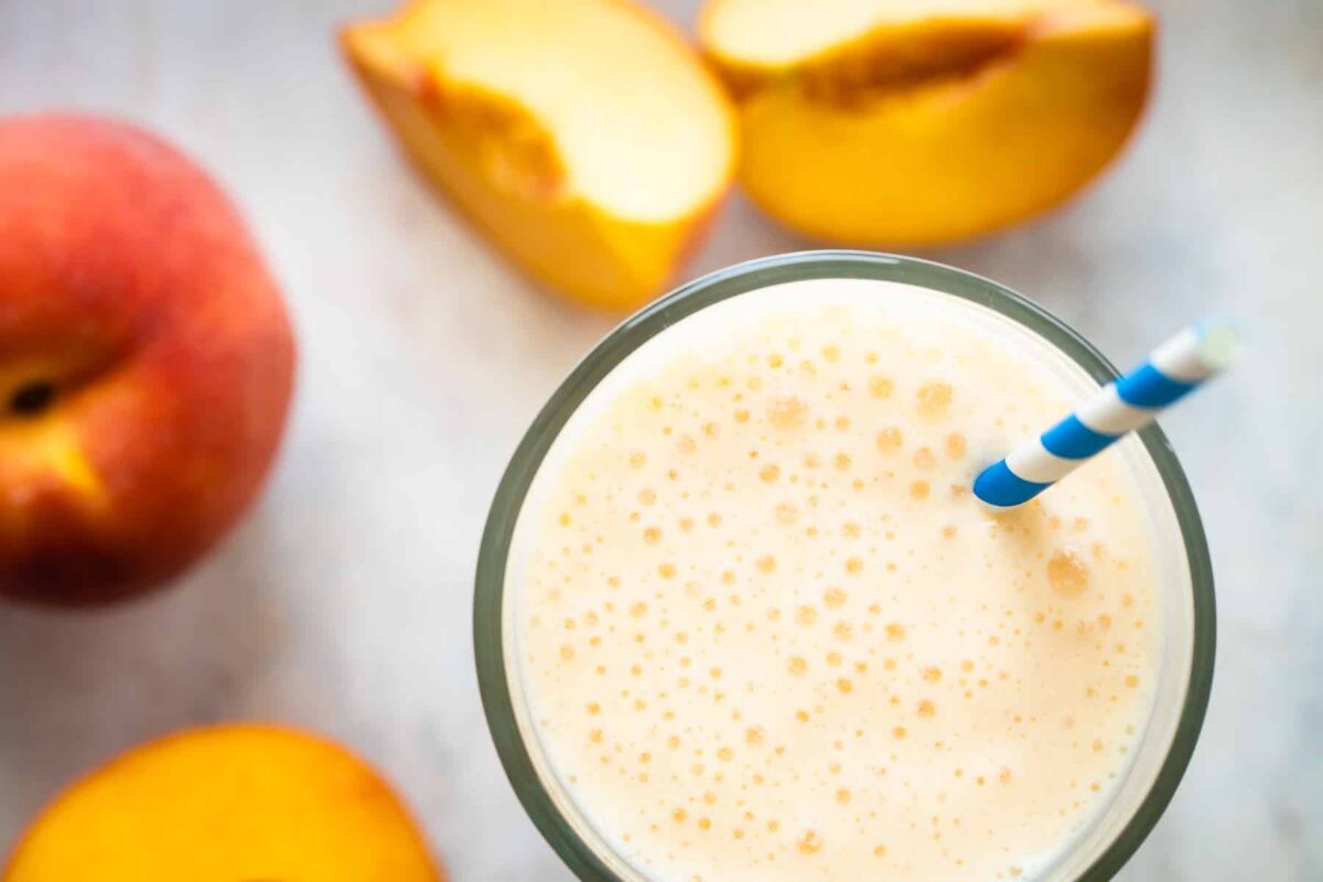 A glass of peach smoothie next to fresh peaches on a counter.