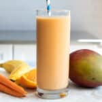 A glass of carrot smoothie next to a mango, carrots, and a banana.