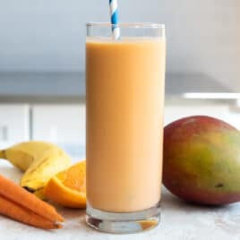 A glass of carrot smoothie next to a mango, carrots, and a banana.