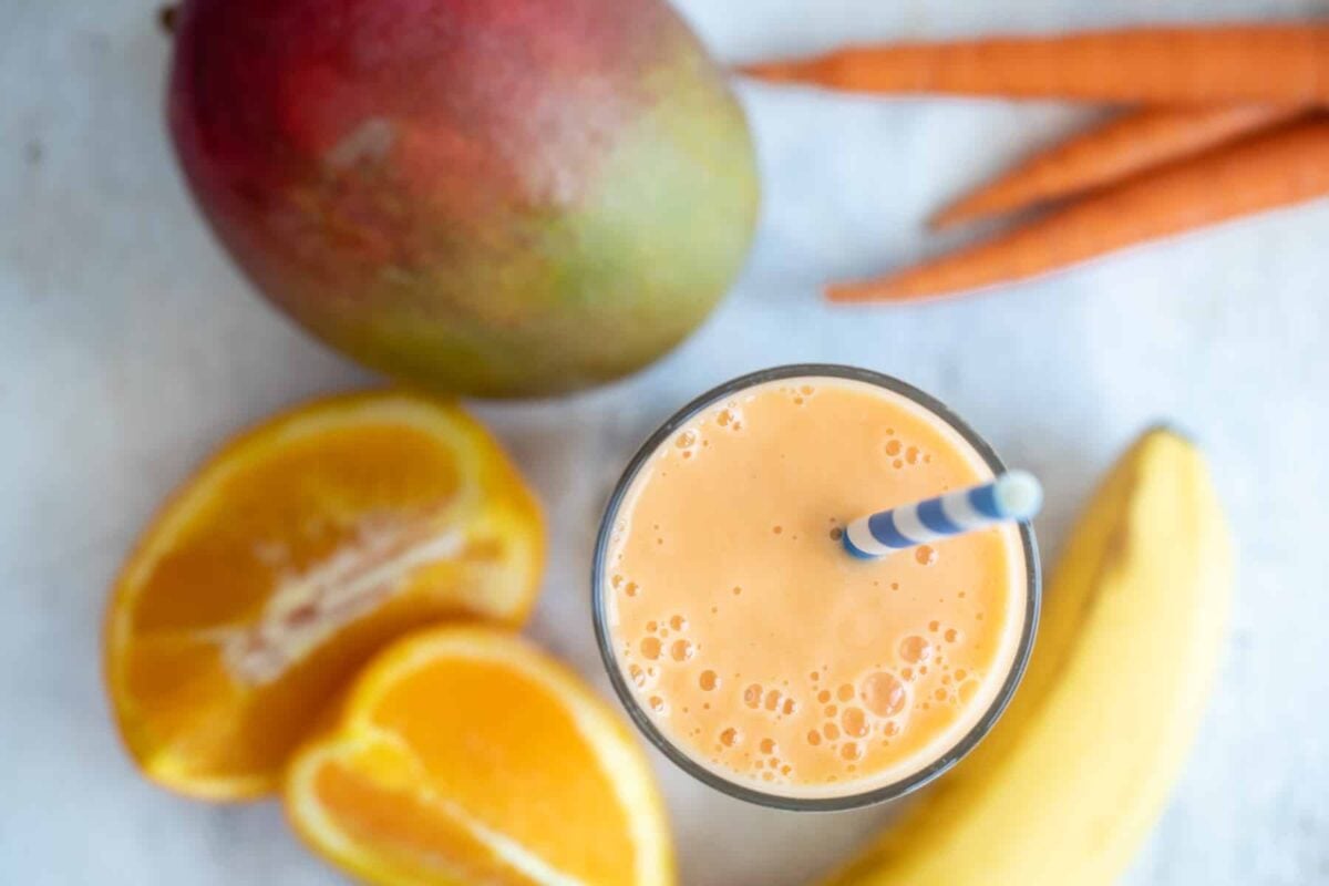 A glass of carrot smoothie next to a mango, carrots, and a banana.