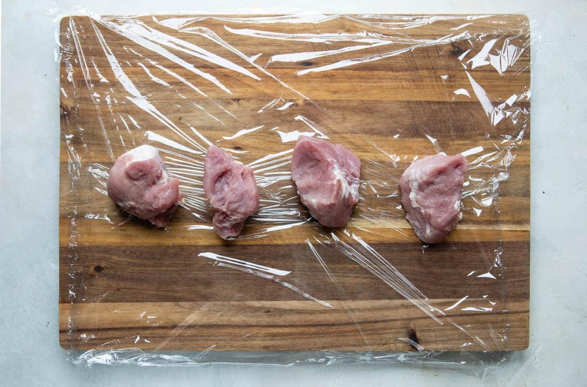 A pork tenderloin cut into 4 pieces, set on a cutting board, covered in plastic.