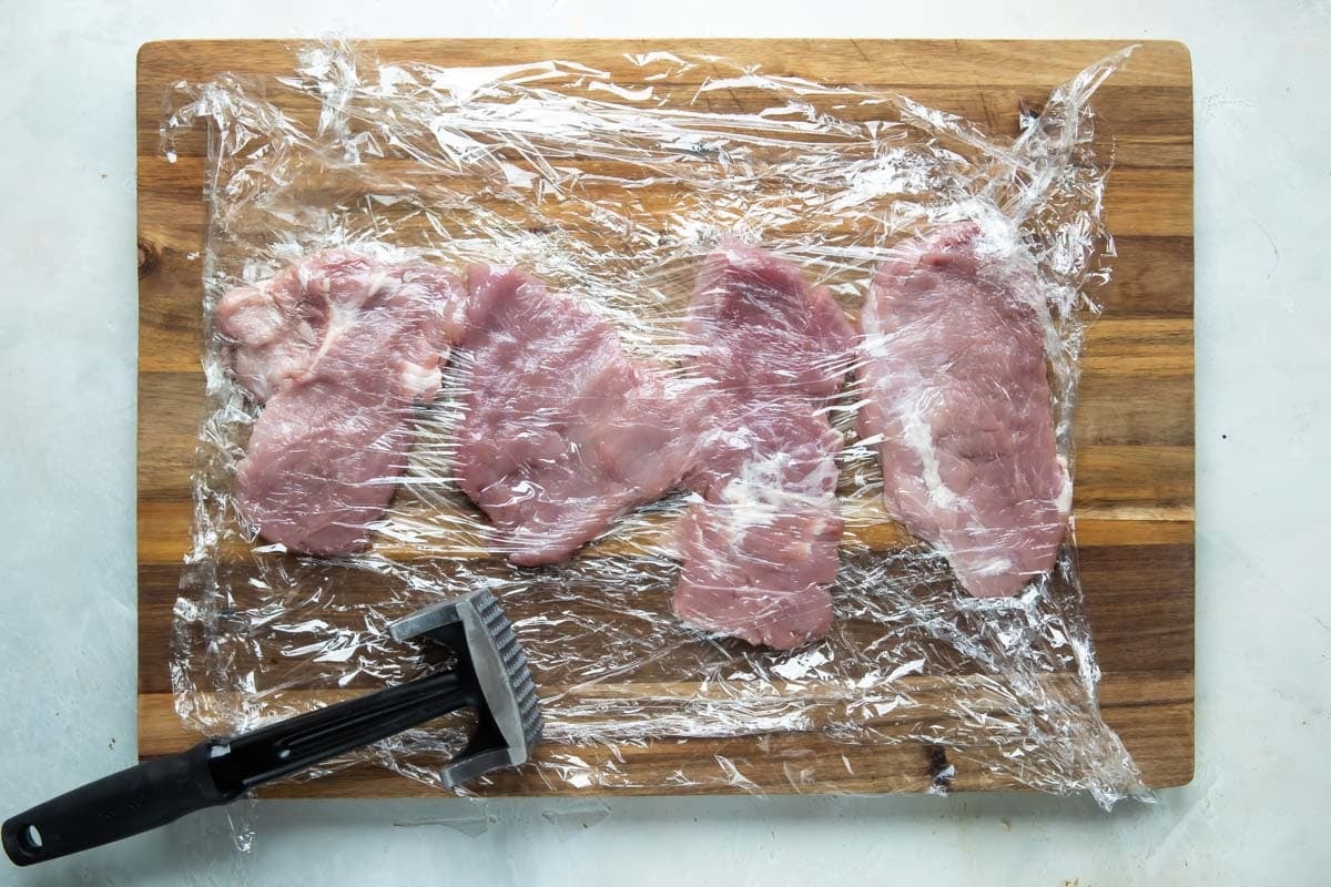 4 pieces of pork tenderloin set on a cutting board, covered in plastic, pounded thin.