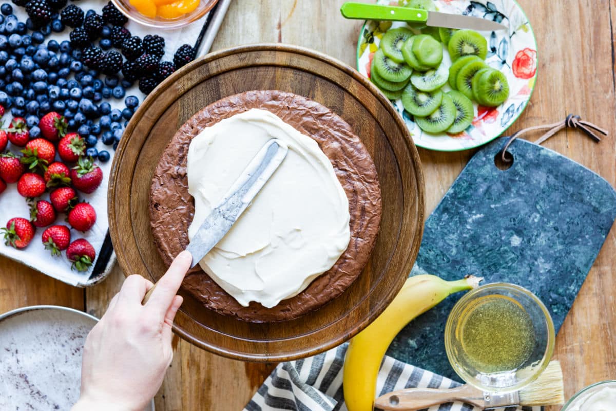 Someone putting cream cheese frosting on top of a round brownie.