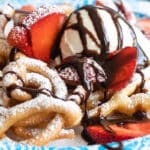 A funnel cake topped with strawberries, ice cream, and chocolate syrup on a paper plate.