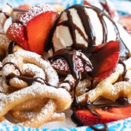A funnel cake topped with strawberries, ice cream, and chocolate syrup on a paper plate.