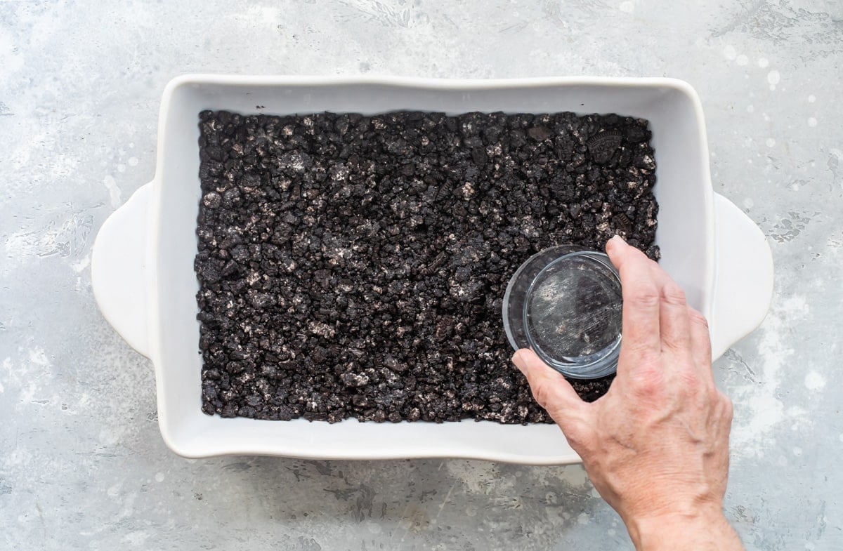Oreo crust being pressed into a white baking dish.