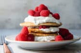 A stack of oatmeal pancakes topped with whipped cream and raspberries on a plate.