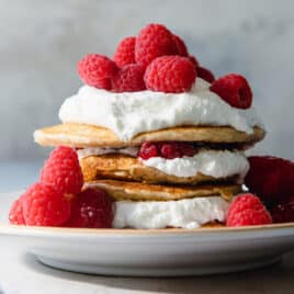 A stack of oatmeal pancakes topped with whipped cream and raspberries on a plate.