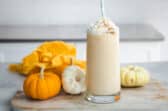 A pumpkin smoothie in a clear glass surrounded by small pumpkins.