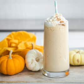 A pumpkin smoothie in a clear glass surrounded by small pumpkins.