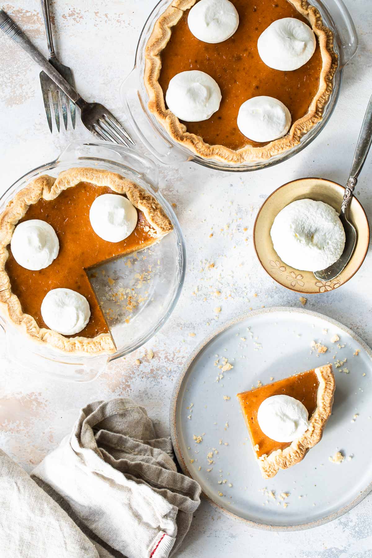 A slice of mini pumpkin pie on a white plate with two mini pumpkin pie and whipped cream behind it.