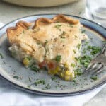 Turkey pot pie in a white pie pan and a piece of turkey pot pie on a blue plate with a fork and a clear glass of water.