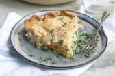 Turkey pot pie in a white pie pan and a piece of turkey pot pie on a blue plate with a fork and a clear glass of water.