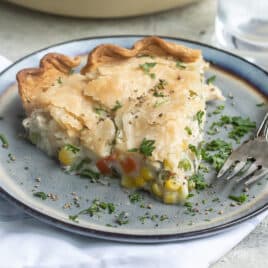 Turkey pot pie in a white pie pan and a piece of turkey pot pie on a blue plate with a fork and a clear glass of water.
