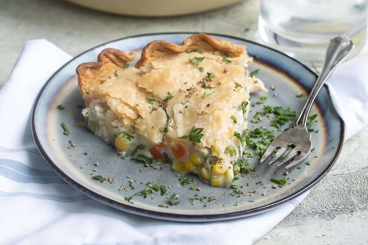 Turkey pot pie in a white pie pan and a piece of turkey pot pie on a blue plate with a fork and a clear glass of water.
