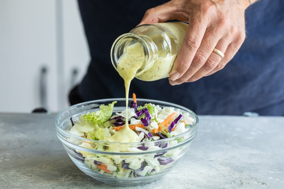 Coleslaw dressing being poured onto a large clear bowl of cabbage.