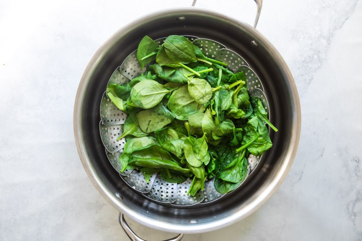 Spinach being steamed in a silver steamer basket inside a silver pan.