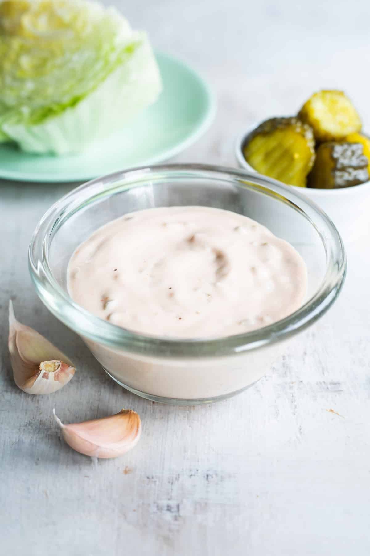 Thousand Island dressing in a clear bowl on a countertop with pickles and lettuce in the background.