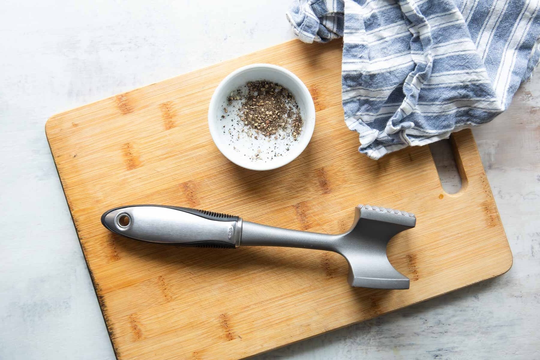 An OXO meat tenderizer on a cutting board next to a bowl of salt and pepper.
