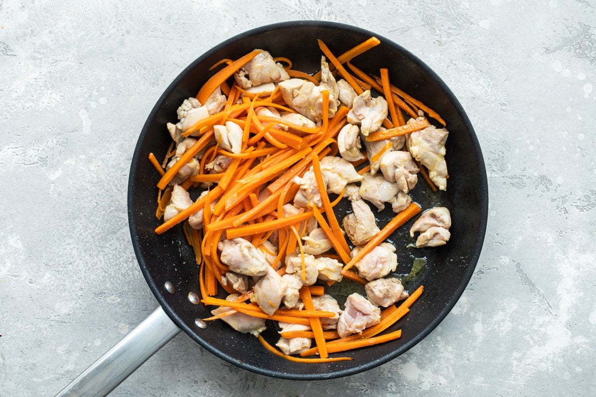 Chunks of chicken and sliced carrots sauteing in a black skillet.