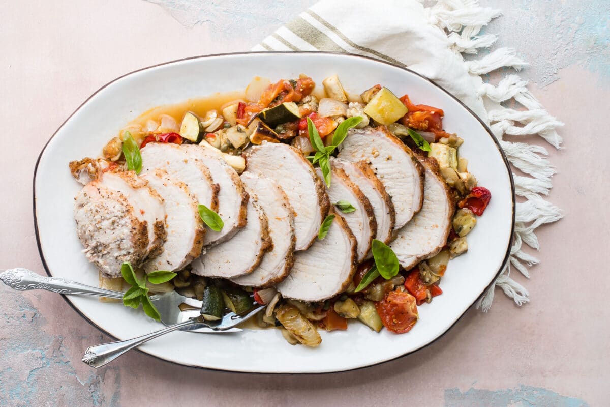 An overhead shot of pork loin with ratatouille on a white platter.