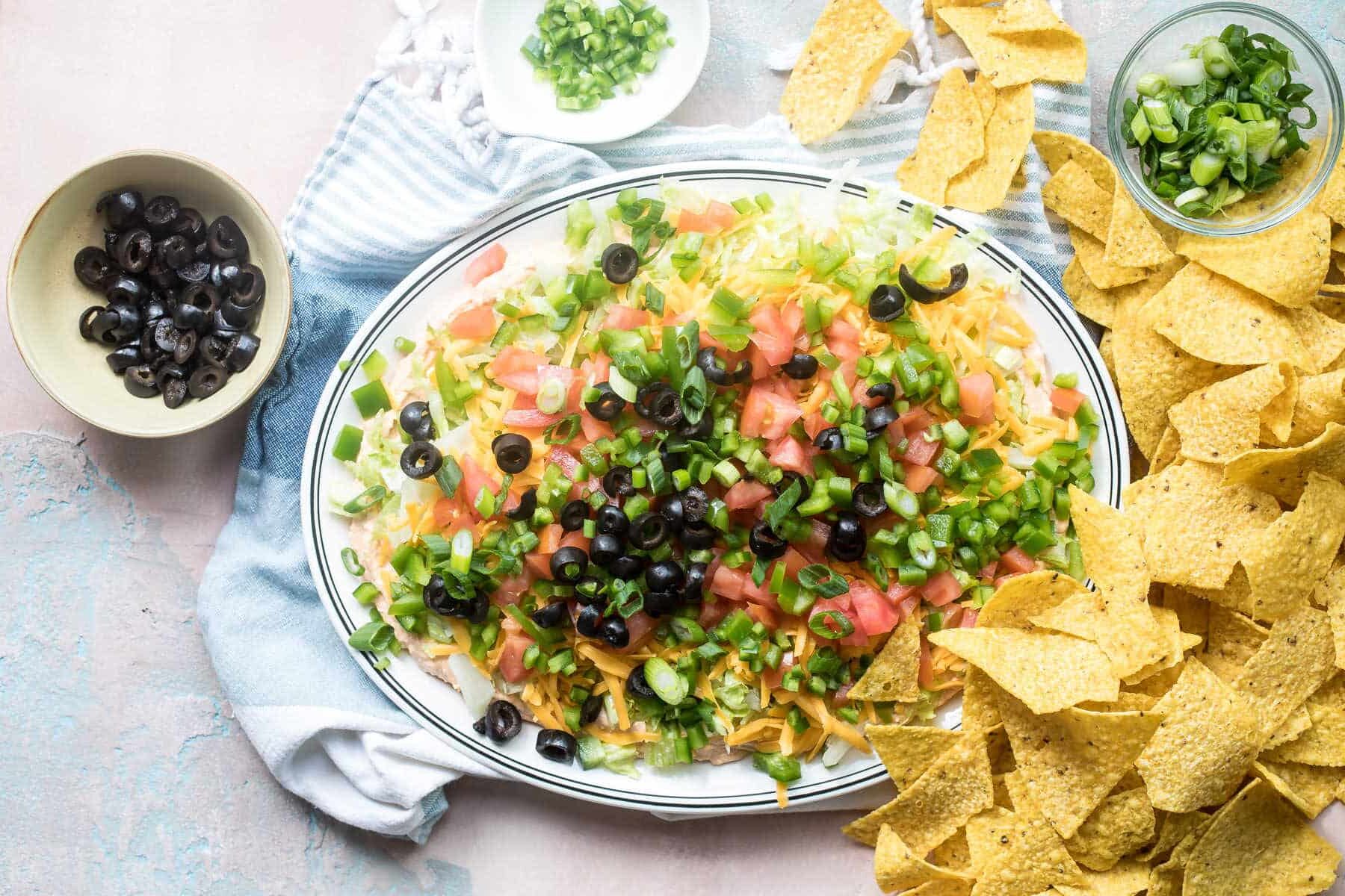 A platter of taco dip surrounded by tortilla chips and small bowls of toppings.
