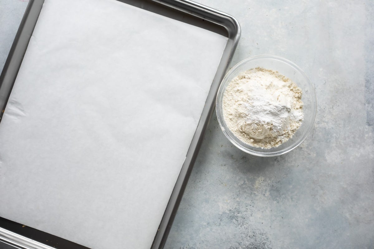 A parchment paper lined baking sheet next to a small bowl of flour.