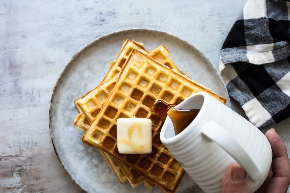 Someone pouring syrup on a stack of waffles on a plate.