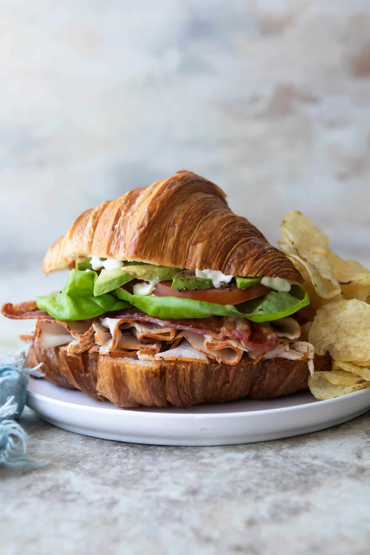 A turkey, bacon and avocado croissant sandwich with potato chips and a pickle on a white plate.