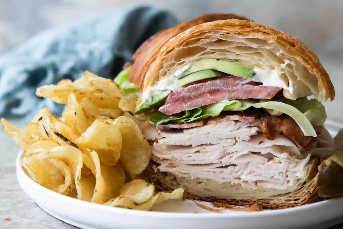 A turkey, bacon and avocado croissant sandwich with potato chips and a pickle on a white plate.