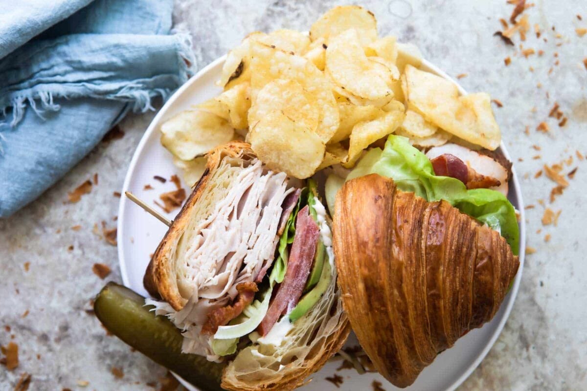 A turkey, bacon and avocado croissant sandwich with potato chips and a pickle on a white plate.