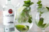 Two mojito cocktails in glasses with a bottle of white rum in the background.
