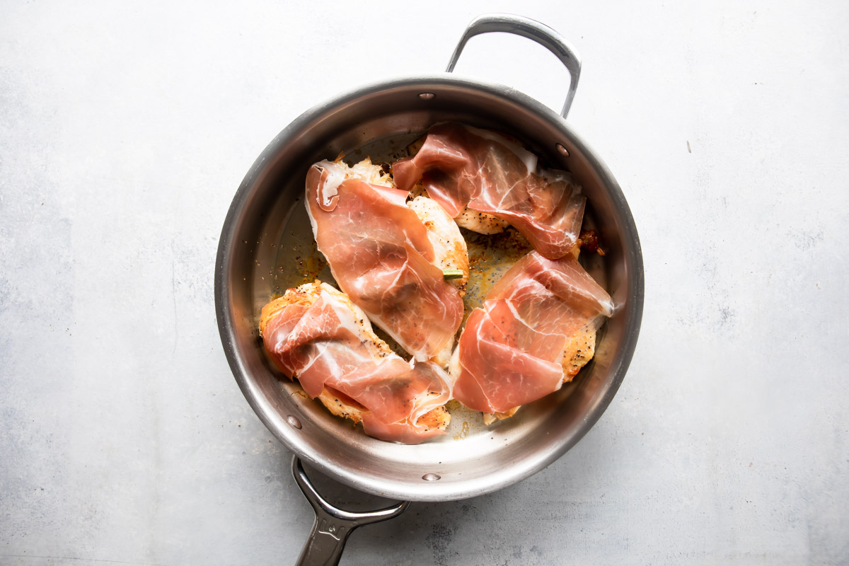 Chicken breasts in a skillet topped with prosciutto.