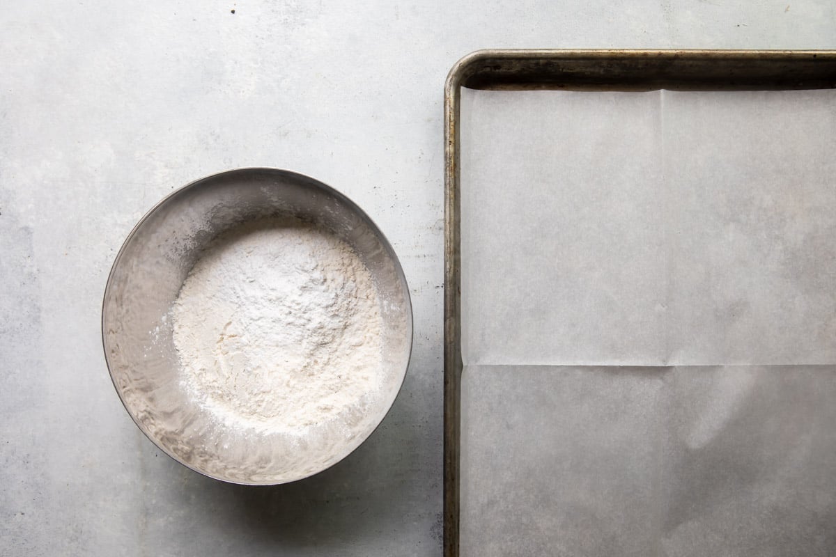 A bowl of flour next to a parchment paper lined baking sheet.
