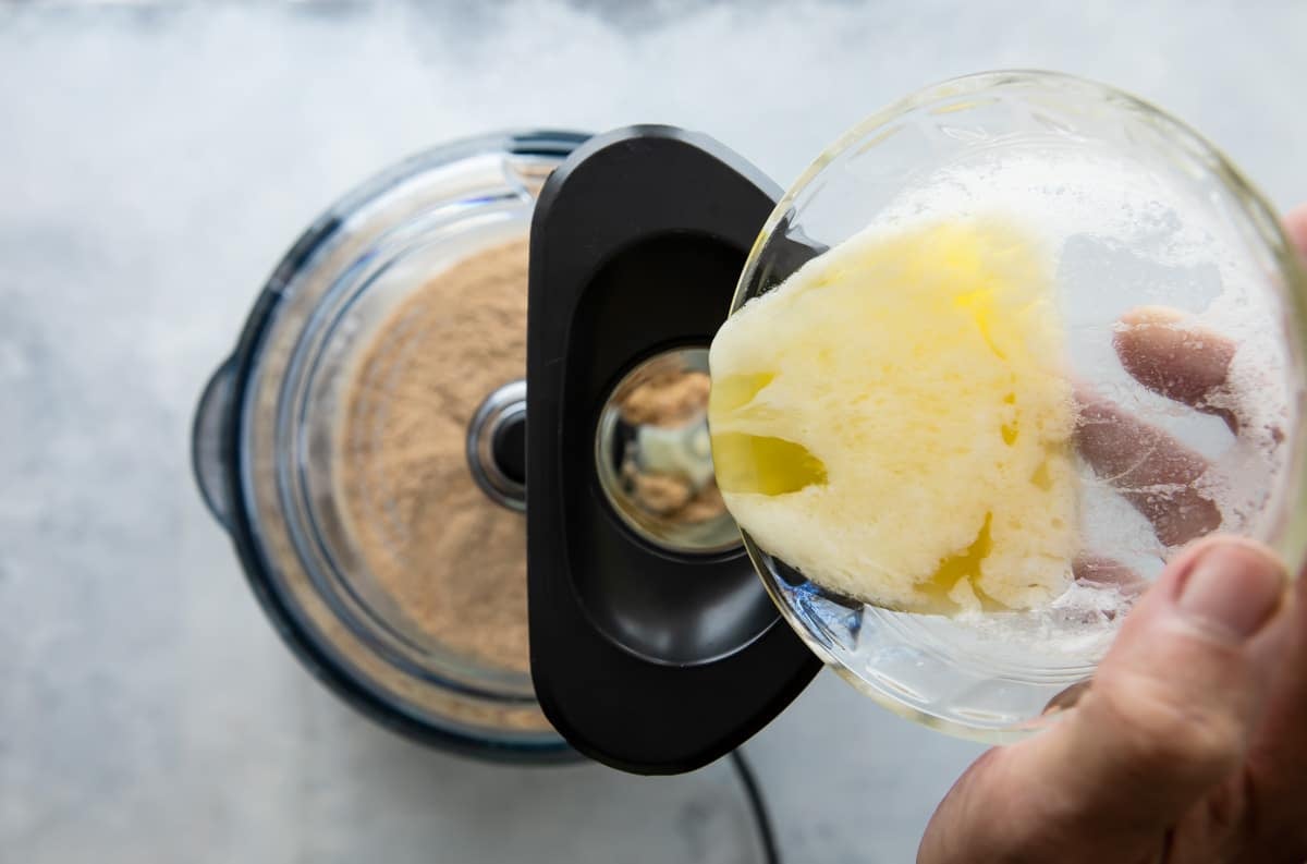 Melted butter being added to graham cracker crumbs in a food processor.
