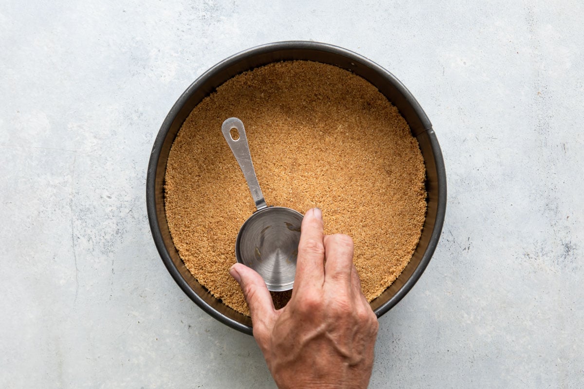Someone flattening out a graham cracker crust with the bottom of a measuring cup.