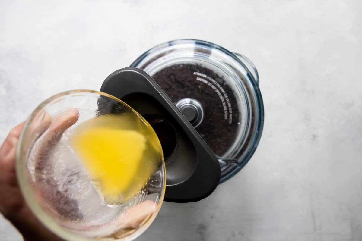 Someone pouring melted butter into a food processor of Oreo crumbs.