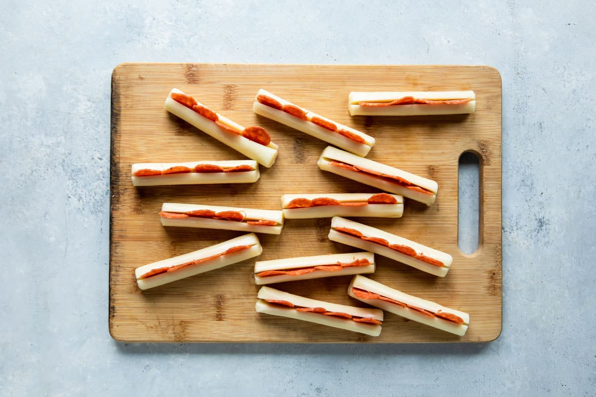 Twelve string cheese pieces on a wooden cutting board with pepperoni wedged in them.