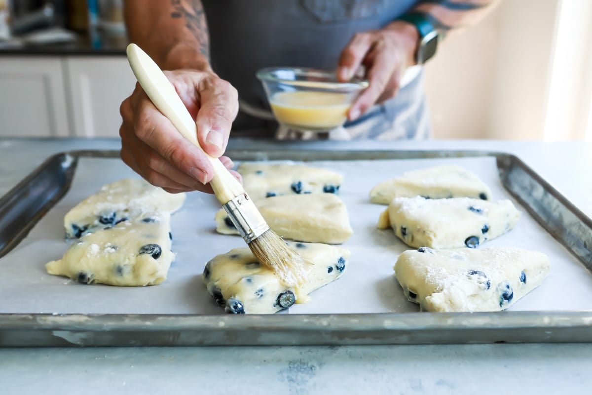 Someone brushing melted butter on blueberry scone dough triangles.