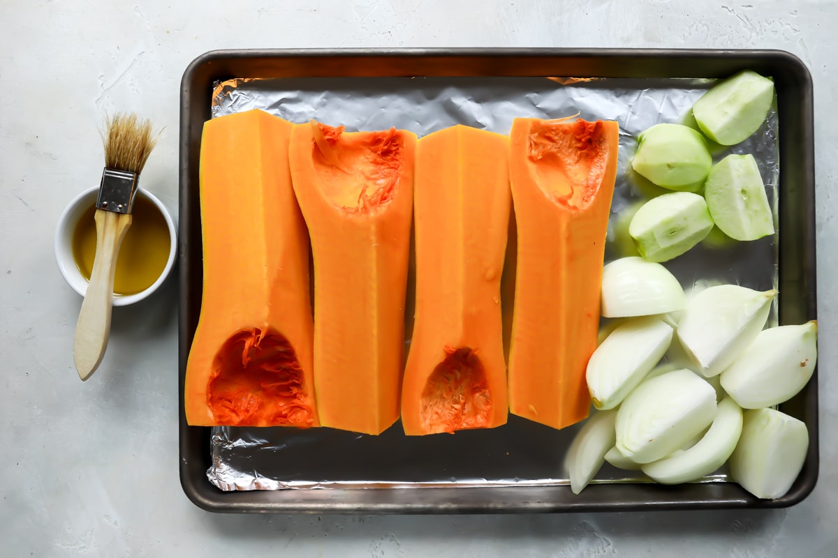 baking sheet with butternut squash quarters, onion quarters, and apple quarters next to small bowl with oil honey mixture and brush.