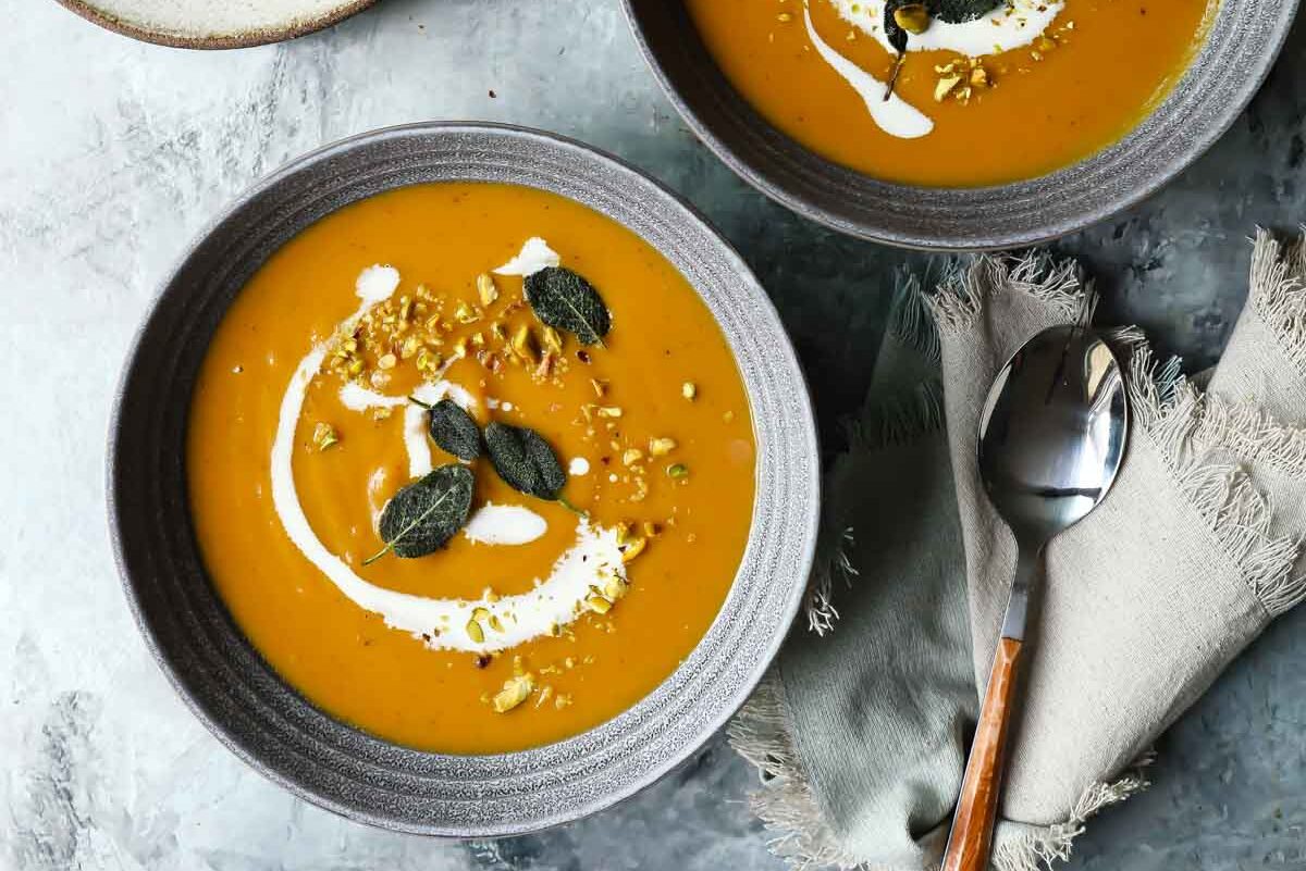 bowls of butternut squash soup with swirl of half and half, fried sage leaves, and chopped pistachios. 