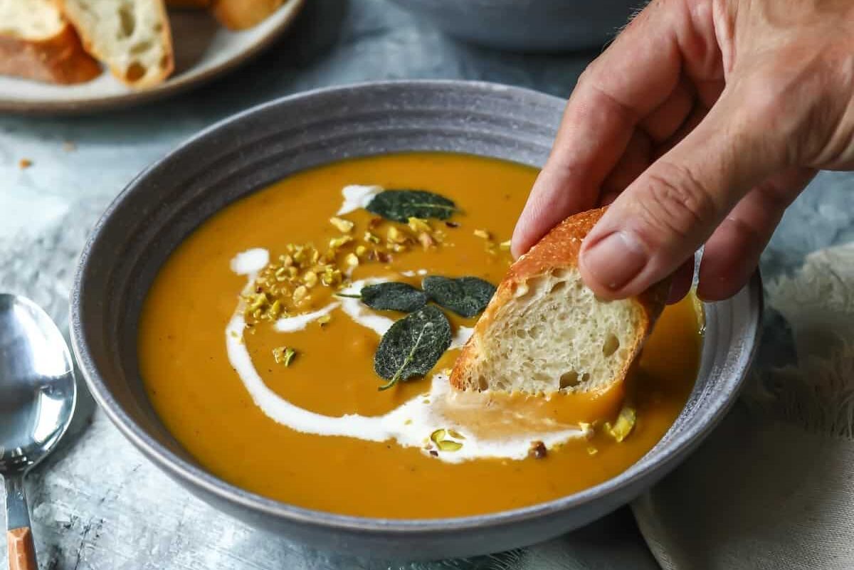 hand dipping crusty bread into bowl of butternut squash soup. 